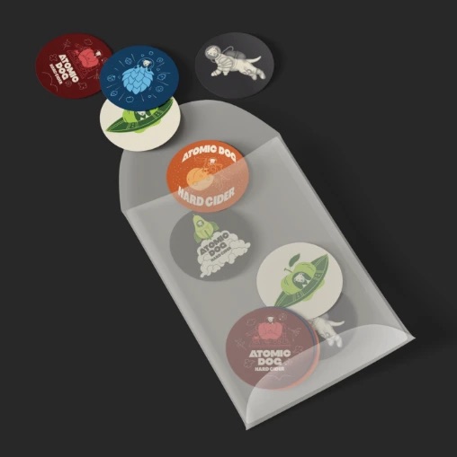 Atomic Dog Hard Cider branded stickers and coasters displayed in a translucent pouch, featuring colorful space-themed cider artwork and logo designs.