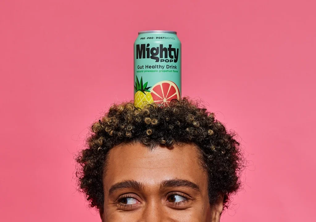 Mighty Pop gut-healthy soda can balanced on a person’s head against a pink background, representing playful consumer goods branding by Agency Squid.